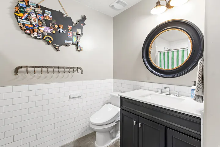 Bathroom featuring a white subway tile wainscoting, a dark vanity with a white countertop, an oval mirror, and a two-light fixture