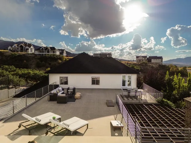 Large exterior patio with concrete and wood-decked areas, featuring a black shingle roof, white exterior walls, and modern metal railings