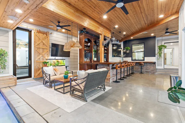 Expansive covered patio featuring a wood-panel ceiling, built-in bar with brick accents, polished concrete flooring, and a sliding barn door