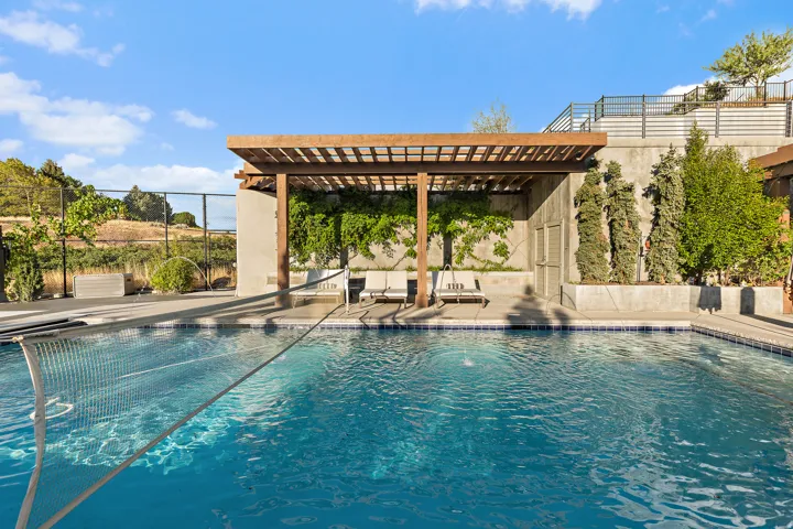 Swimming pool with a perimeter of blue mosaic tile, concrete decking, and a wood pergola