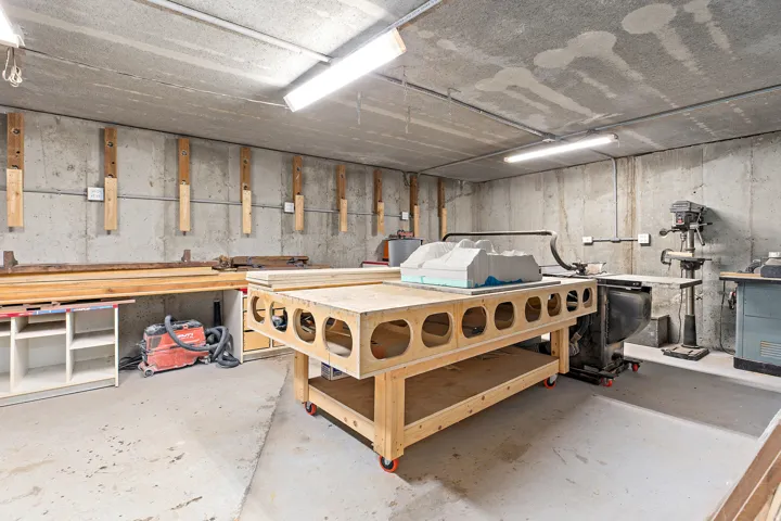 Concrete walls and ceiling, fluorescent lighting, concrete flooring, built-in shelving, and a mobile wooden workbench