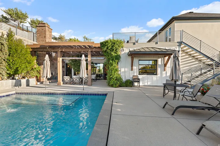 Expansive outdoor living area featuring a rectangular swimming pool with tiled coping, a wood-beam pergola, a brick chimney, and concrete patio decking