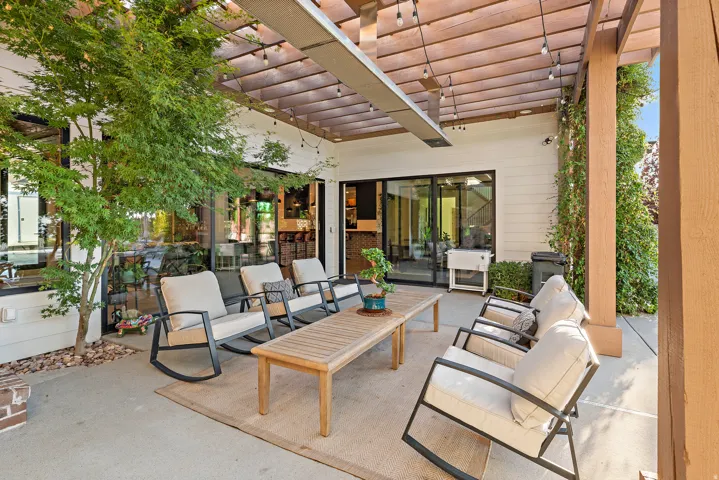 Spacious outdoor patio featuring a wood pergola, string lighting, a wall-mounted heater, mature landscaping, and sliding glass doors