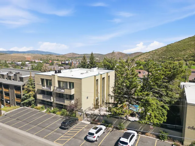 Multi-story building with balconies, surrounding mature trees, and an asphalt parking area