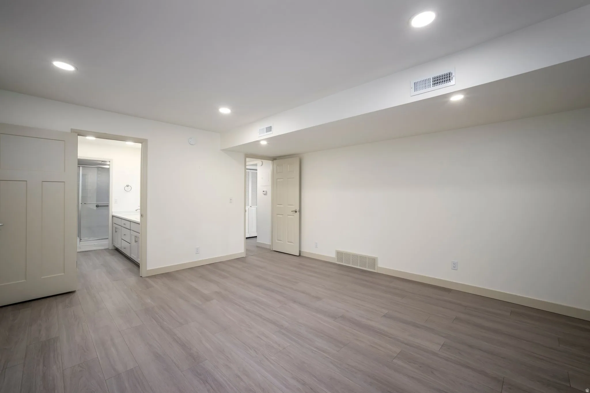 Recessed lighting illuminates wood-finish flooring, white walls, and a visible doorway leading to a bathroom with a glass-enclosed shower and vanity