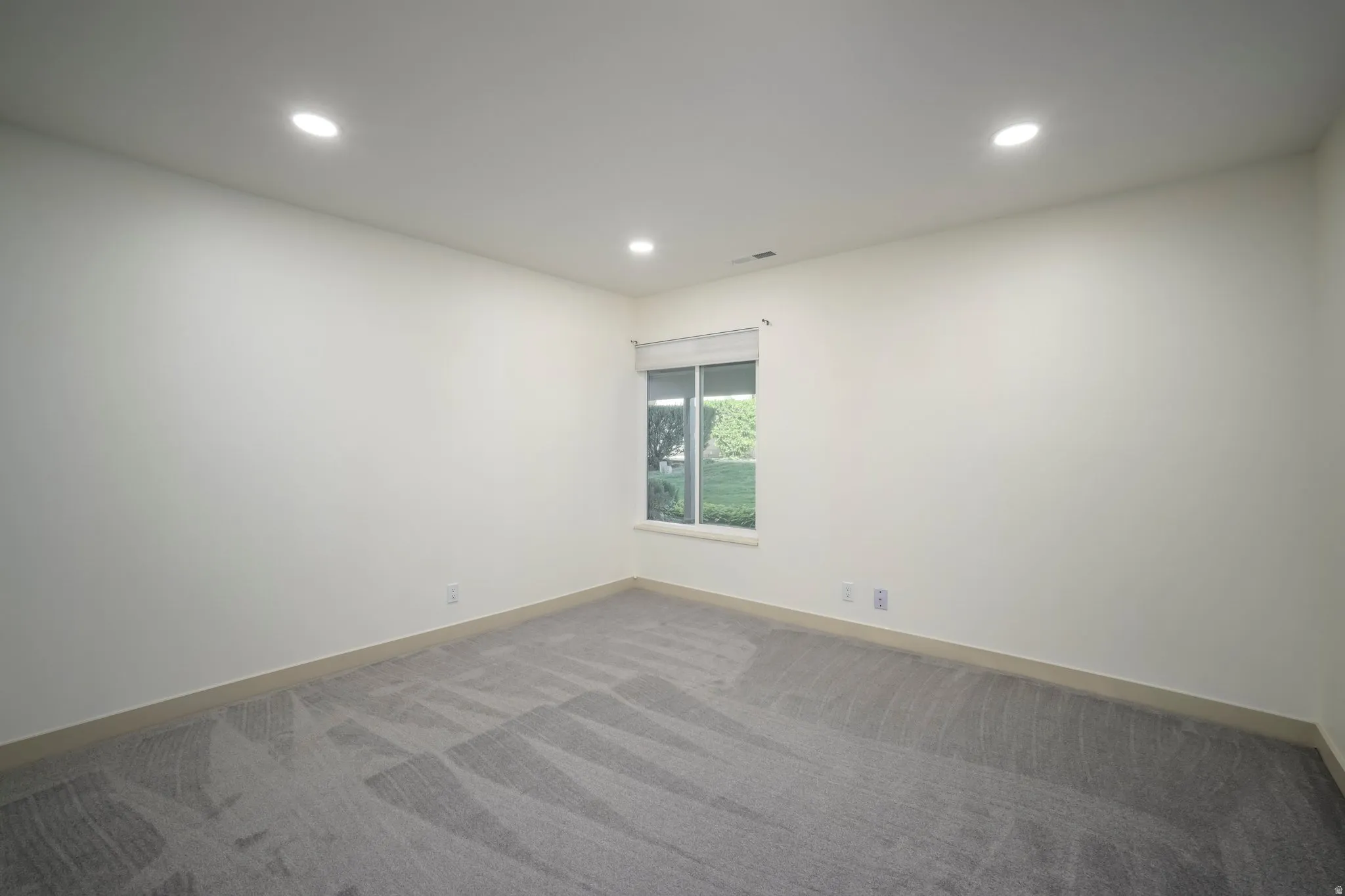 Recessed ceiling lighting, patterned carpet flooring, single window with roller shade, neutral wall tones, and light-toned baseboards