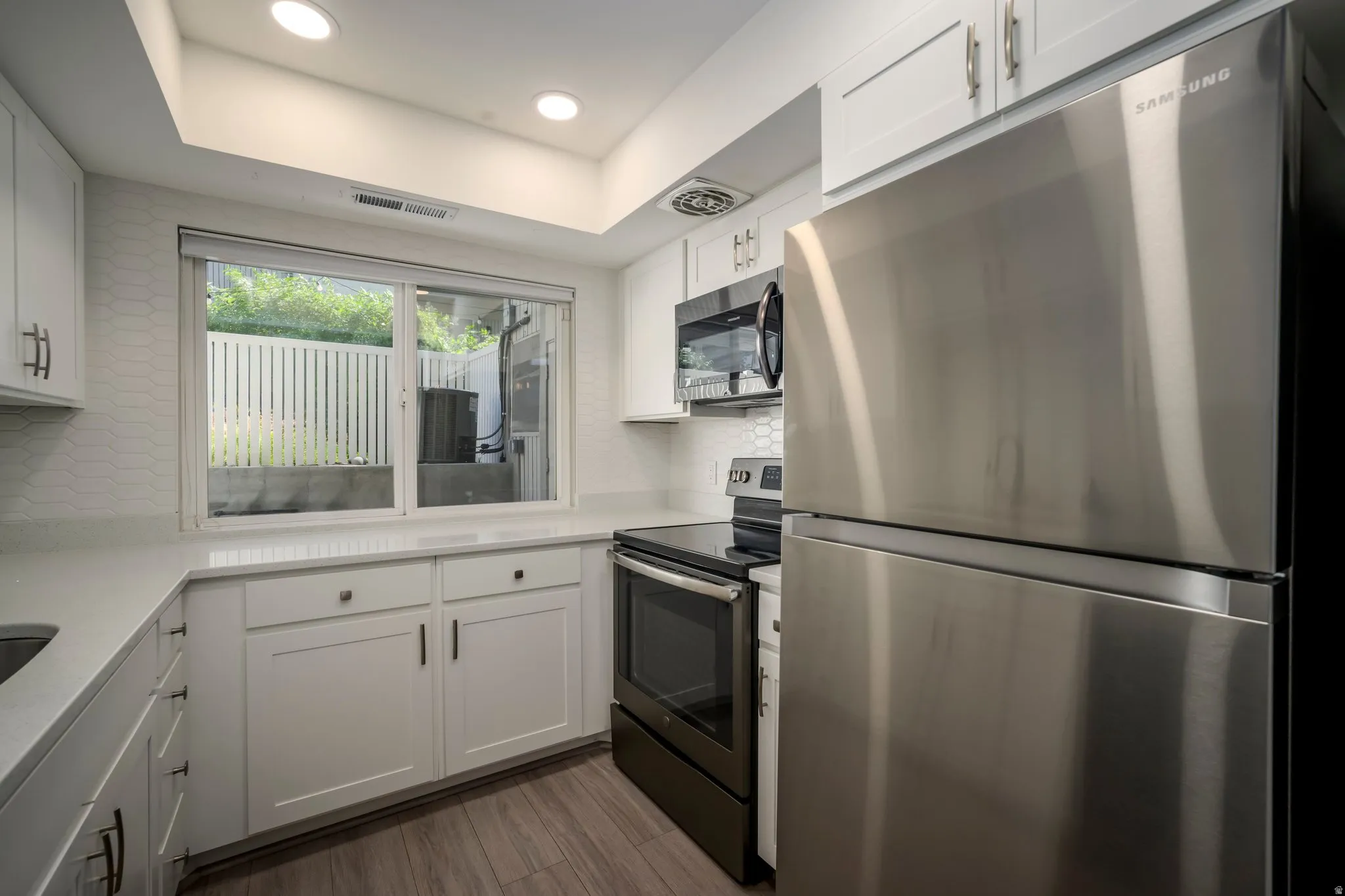 White shaker-style cabinetry, stainless steel appliances, light-toned countertops, contemporary geometric tile backsplash, and wood-finish flooring