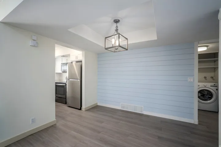 Wood-finish flooring, accent wall with horizontal paneling, recessed ceiling detail, contemporary chandelier, and stainless steel kitchen appliances