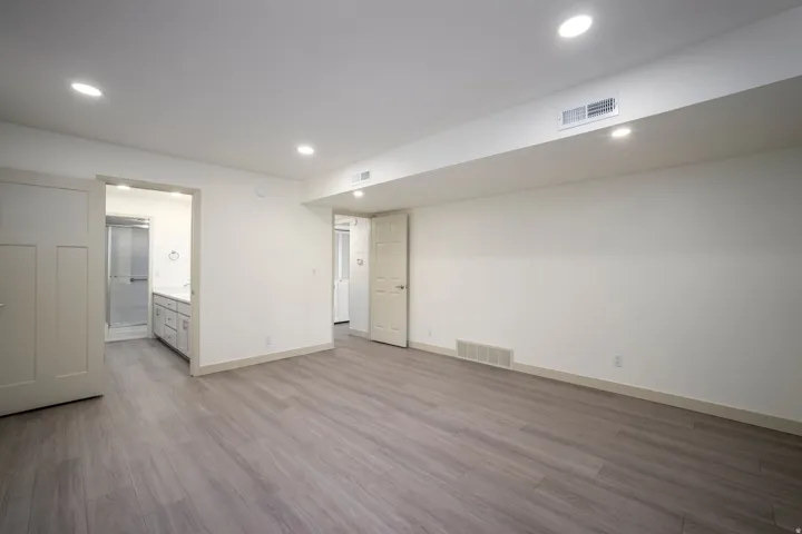 Recessed lighting illuminates wood-finish flooring, white walls, and a visible doorway leading to a bathroom with a glass-enclosed shower and vanity
