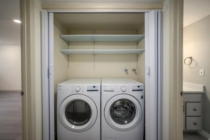 Laundry closet with bi-fold doors, white washer and dryer units, and two wall-mounted shelves