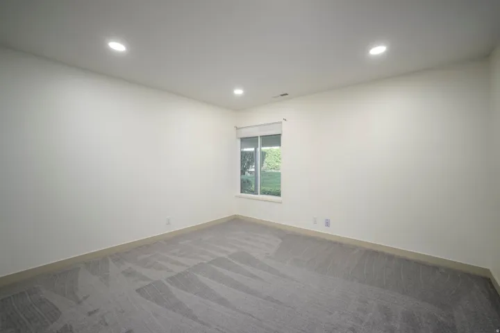Recessed ceiling lighting, patterned carpet flooring, single window with roller shade, neutral wall tones, and light-toned baseboards
