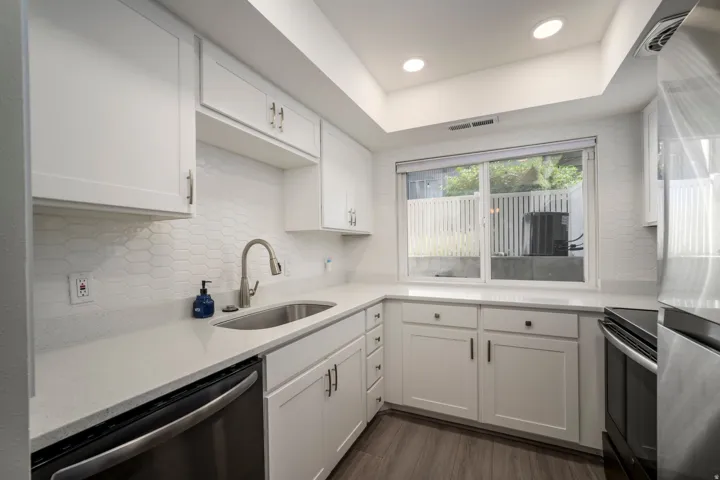 White shaker cabinetry with brushed nickel hardware, white stone countertops, hexagonal tile backsplash, stainless steel sink with gooseneck faucet, and wood-finish flooring