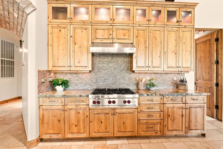 Wood cabinetry with upper glass-front display cabinets, mosaic tile backsplash, and a stainless steel range with matching hood