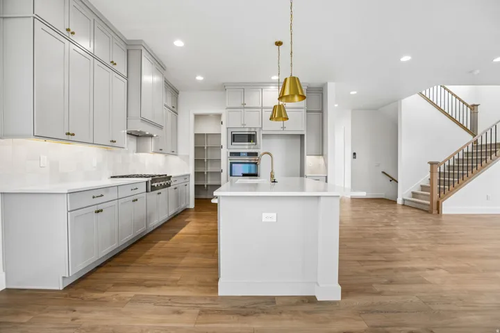 Contemporary kitchen featuring shaker cabinetry, under-cabinet lighting, a central island with a gold-finish faucet, and wood-finish flooring extending to a staircase with a wood handrail and iron balusters