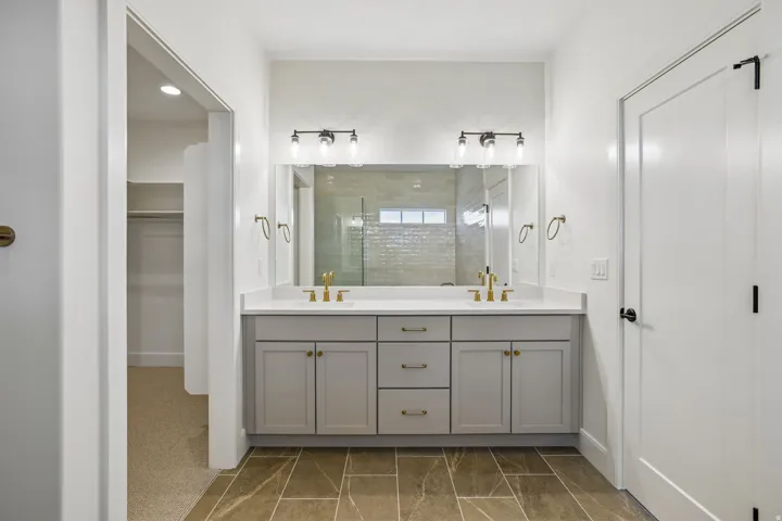 Dual vanity with light gray cabinetry, gold-finish fixtures, and a white countertop, set against a large mirror