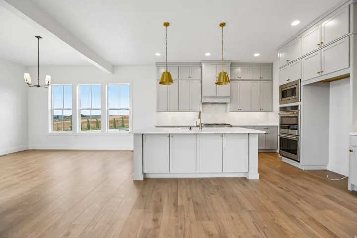 Expansive kitchen island with an undermount sink, light-toned cabinetry, stainless steel built-in appliances, wood-finish flooring, and multiple large windows