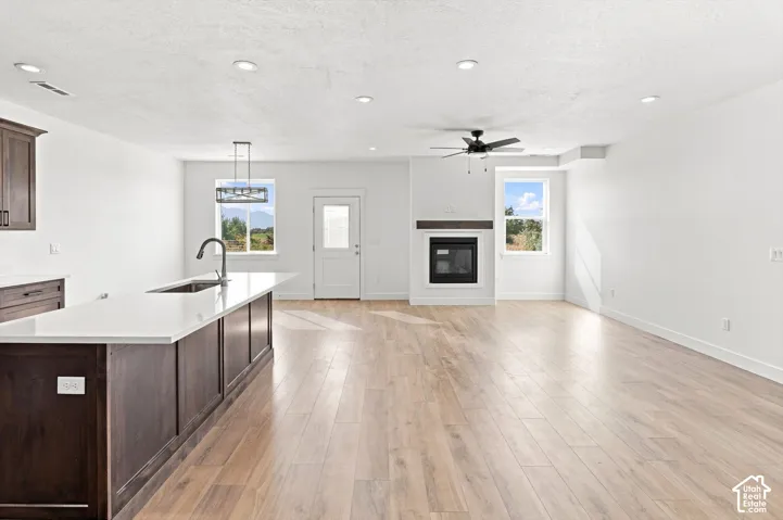 Open-concept living area with wood-finish flooring, a kitchen island with a sink and white countertop, a built-in fireplace with a wood mantel, a ceiling fan, and recessed lighting