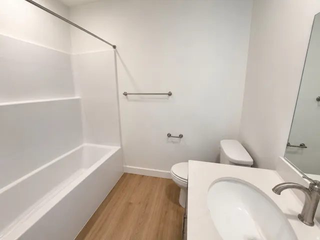 White bathtub with shower surround, wood-finish flooring, white vanity with integrated sink, brushed nickel faucet, and a wall-mounted mirror
