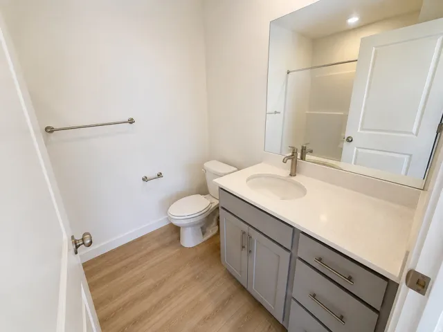 Wood-finish flooring, single vanity with light gray cabinetry, white countertop, undermount sink, and brushed nickel fixtures