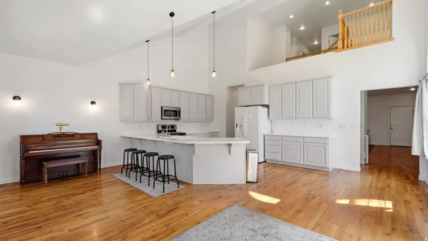 Open-concept living space with high ceilings, wood-finish flooring, a kitchen island with seating, a full suite of kitchen appliances, and a lofted area with a wood railing