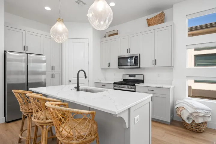 Modern kitchen featuring stainless steel appliances, light gray cabinetry, a central island with a built-in sink, pendant lighting, and multiple windows