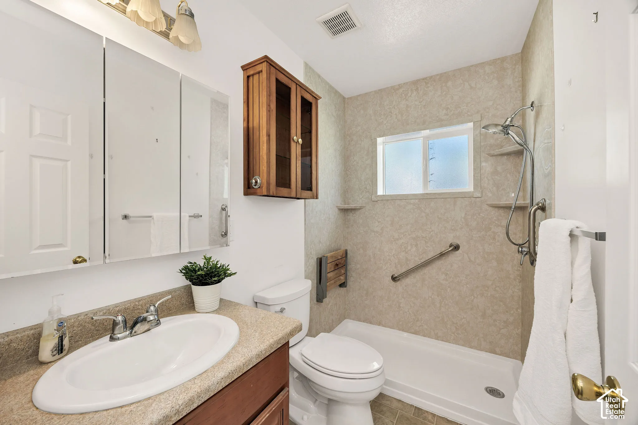 Vanity with an oval sink and speckled countertop, wood-finish cabinetry, triple mirrored medicine cabinet, and a shower with a built-in bench