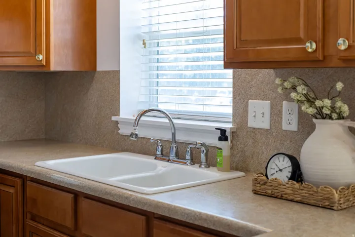 Wood-finish cabinetry, double basin sink, chrome faucet, window with blinds, and light-colored countertop