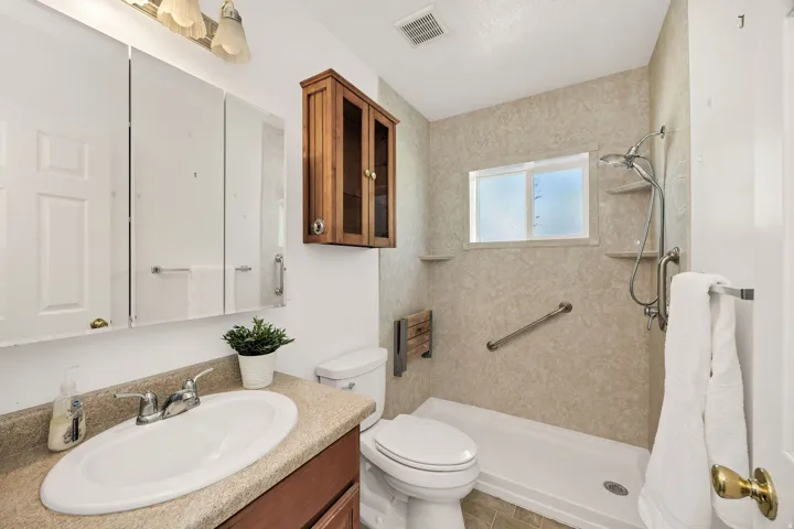 Vanity with an oval sink and speckled countertop, wood-finish cabinetry, triple mirrored medicine cabinet, and a shower with a built-in bench