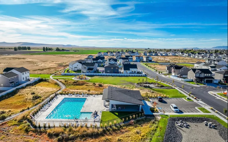 Community swimming pool with surrounding lounge chairs, modern clubhouse building, and winding pedestrian paths, bordered by residential properties and open fields