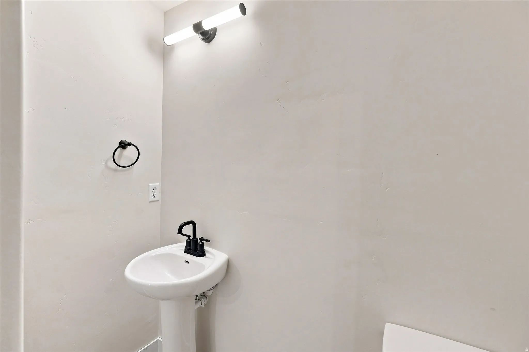 Pedestal sink with a matte black faucet, wall-mounted light fixture, and a black towel ring