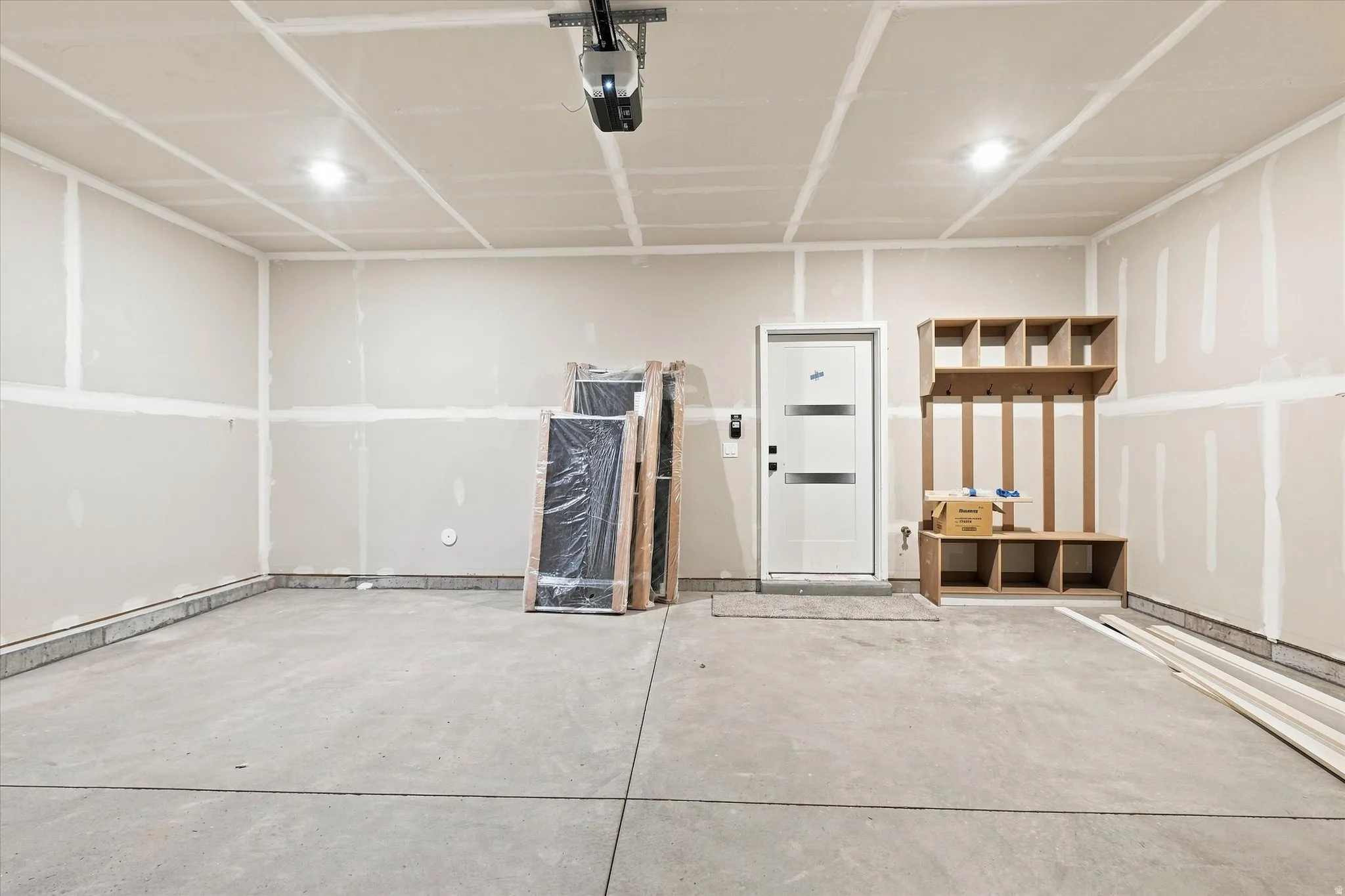 Concrete flooring, ceiling-mounted lighting, garage door opener, exterior door with horizontal paneling, built-in storage unit with shelving and hooks