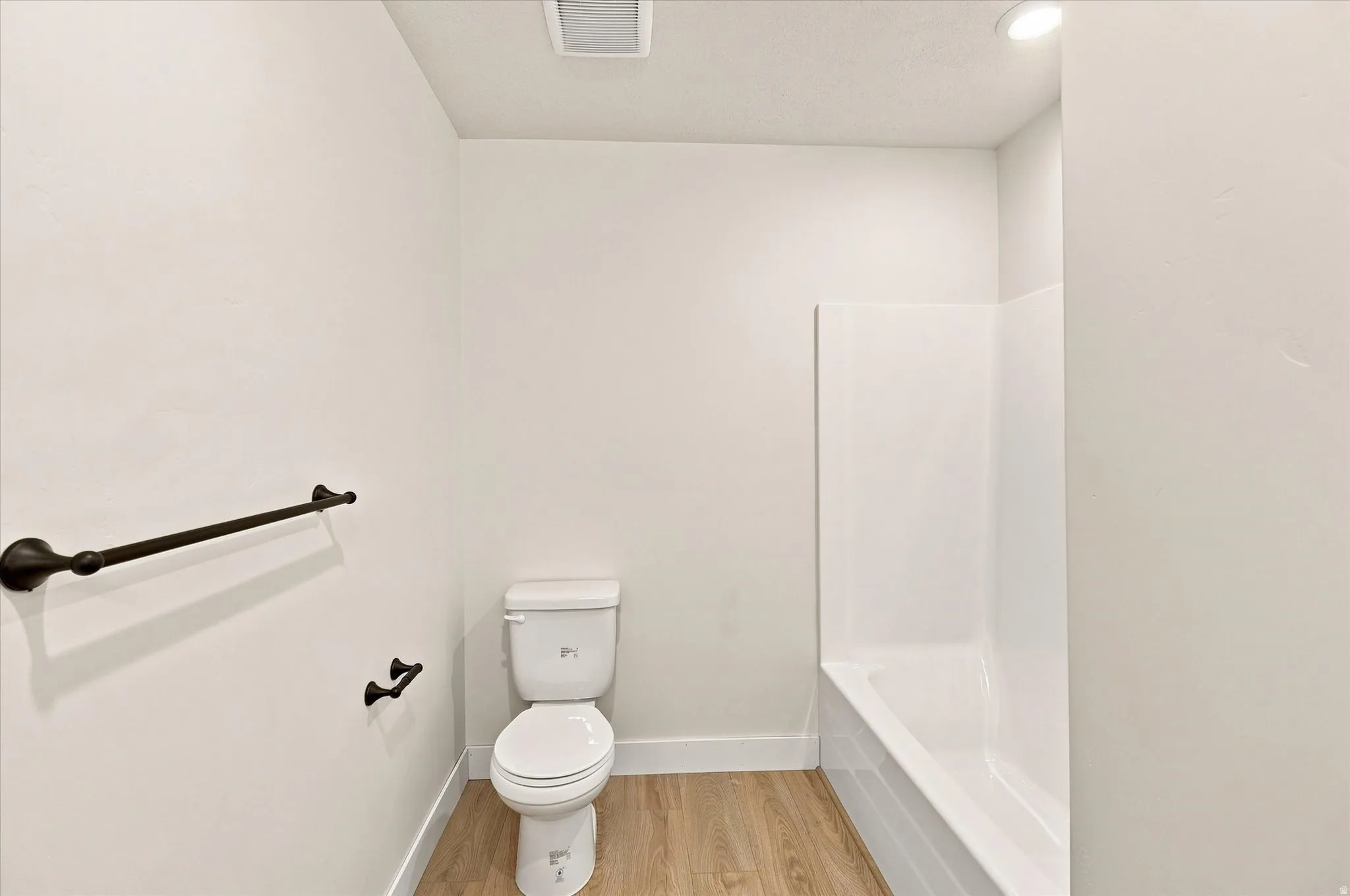 Bathroom featuring a white toilet, white bathtub with wall surround, wood-finish flooring, and dark-finish towel bars