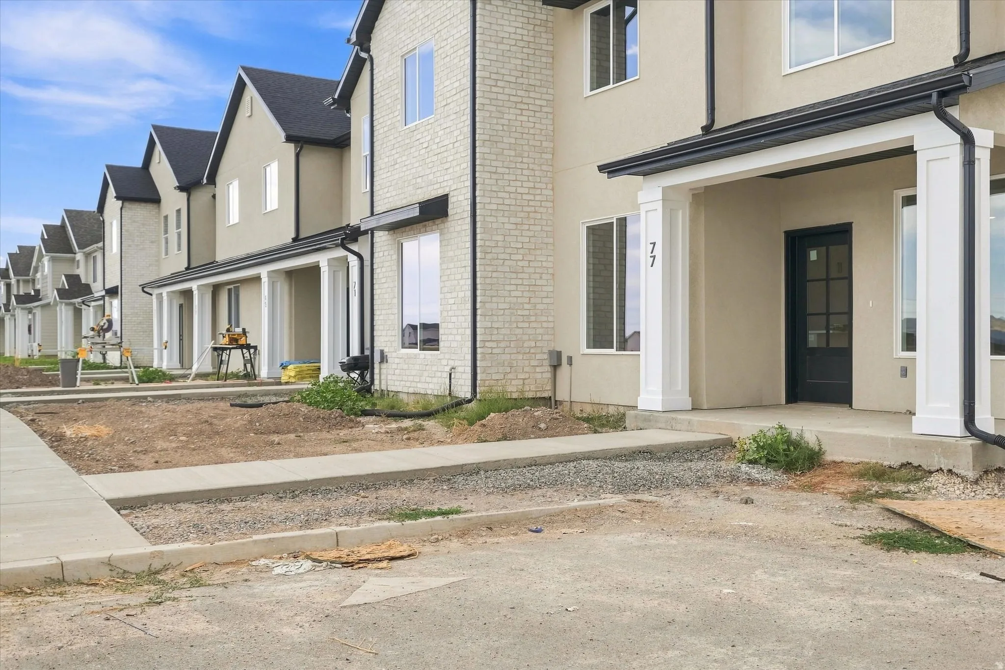 Contemporary townhome featuring a neutral stucco exterior, brick accent details, a black multi-pane entry door, and white columned porch