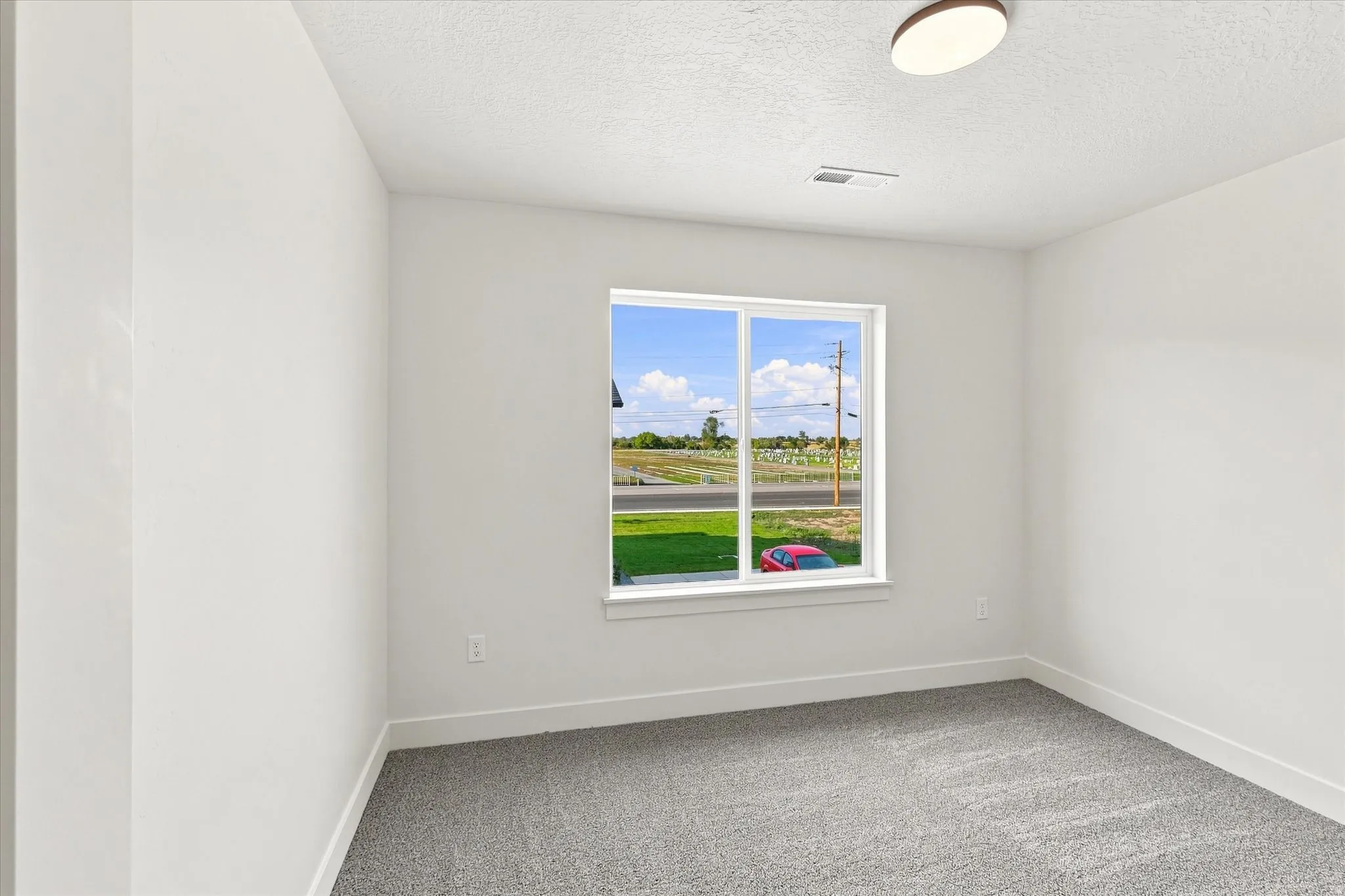 White walls, gray carpet flooring, and a single window with white trim, complemented by a flush-mount ceiling light fixture
