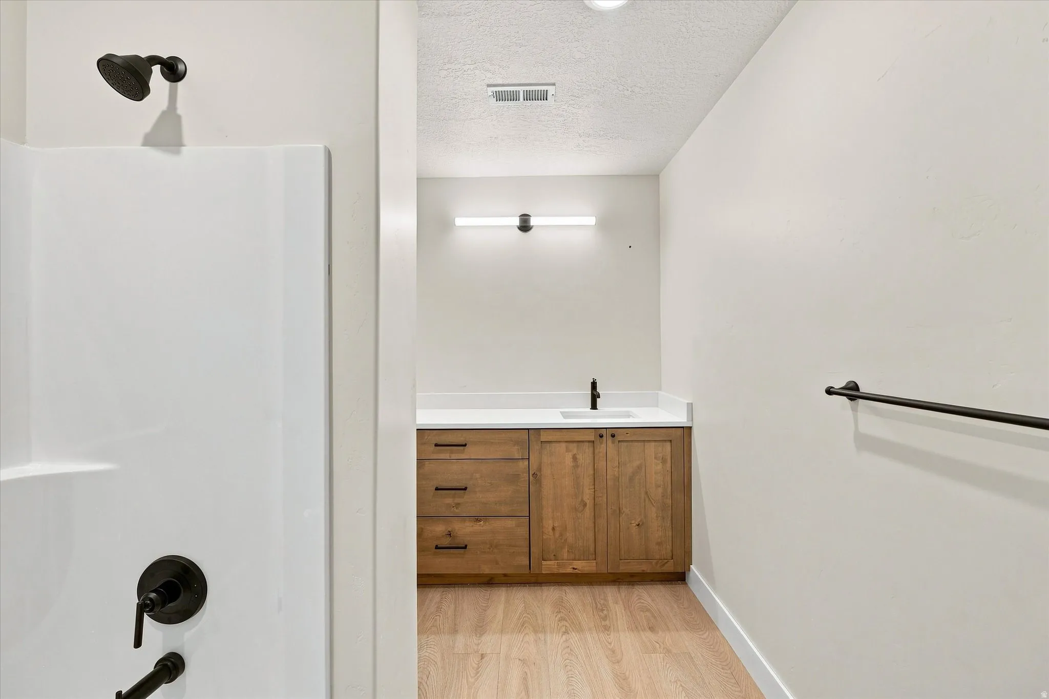 White shower surround with a matte black shower head and controls, wood-finish flooring, a wood-finish vanity with a white countertop and dark-finish sink faucet, and a wall-mounted light fixture
