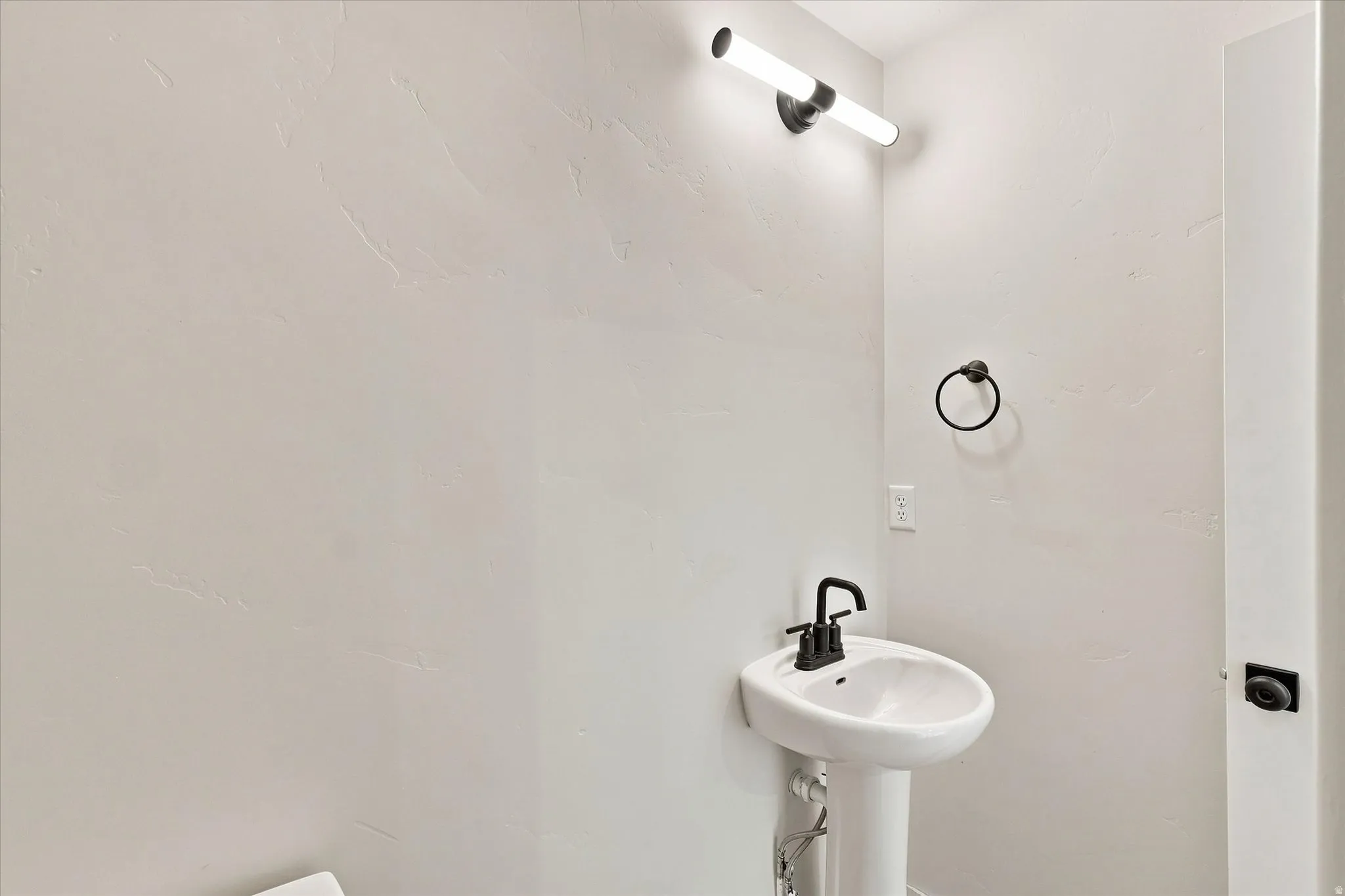 Pedestal sink with a matte black faucet, cylindrical vanity light, and a black circular towel holder