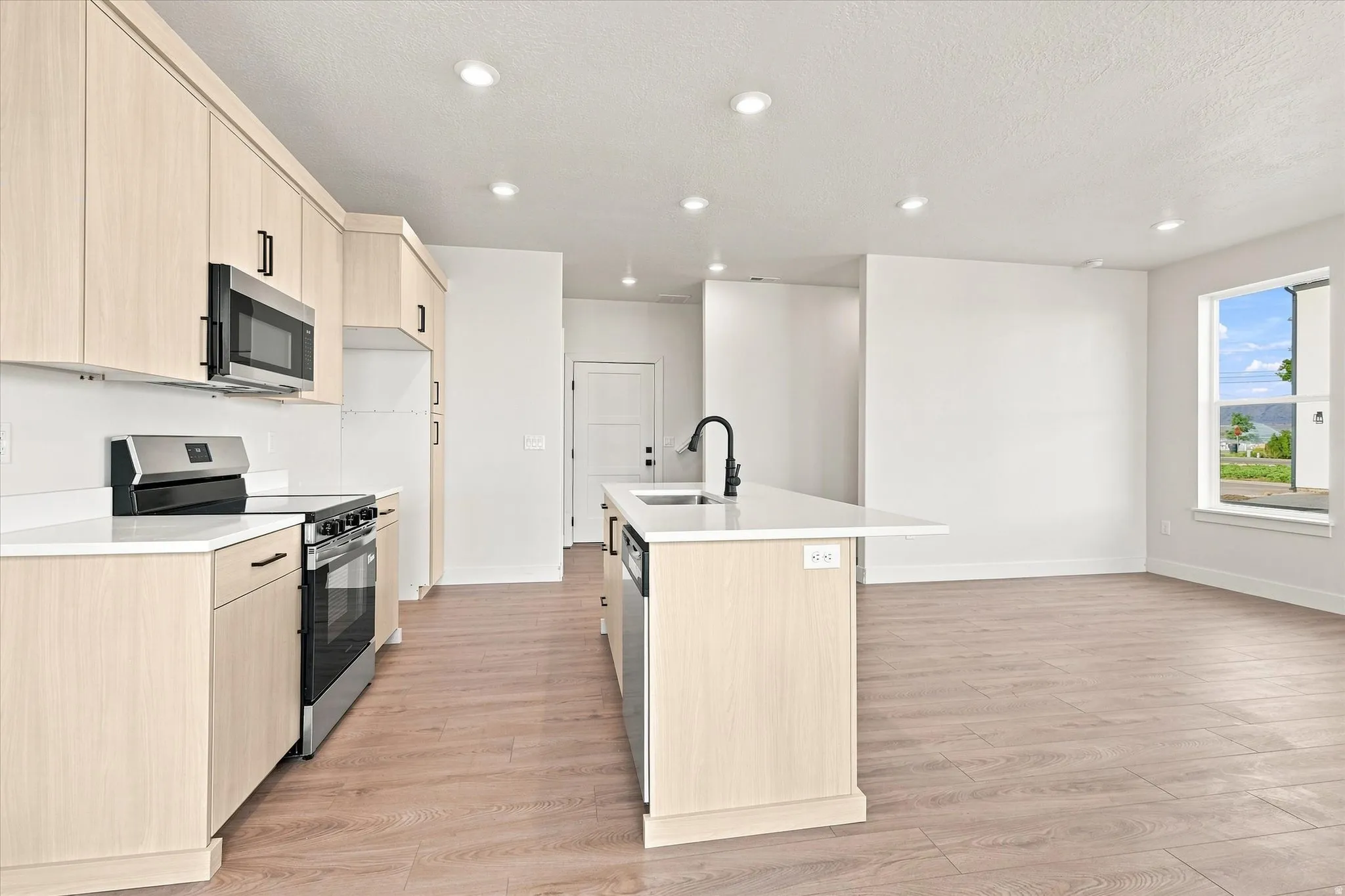 Open-concept kitchen featuring light-toned cabinetry, stainless steel appliances, a center island with a sink, and wood-finish flooring