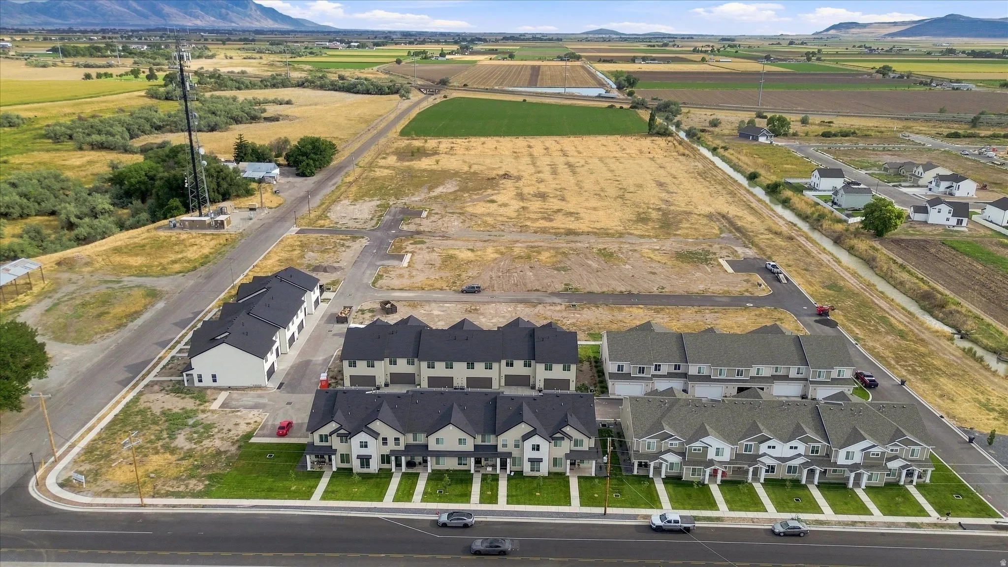 Newly constructed residential development featuring multiple townhome buildings, landscaped common areas, and surrounding open fields with distant mountains