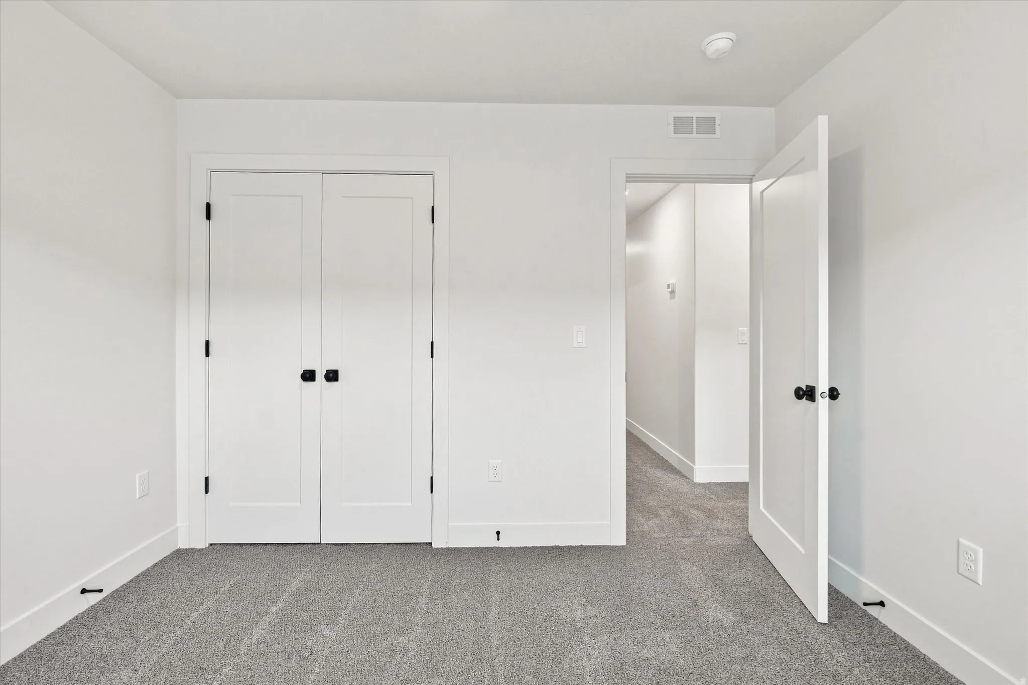 Gray carpet flooring, white walls, white bi-fold closet doors with black hardware, white solid core door with black hardware