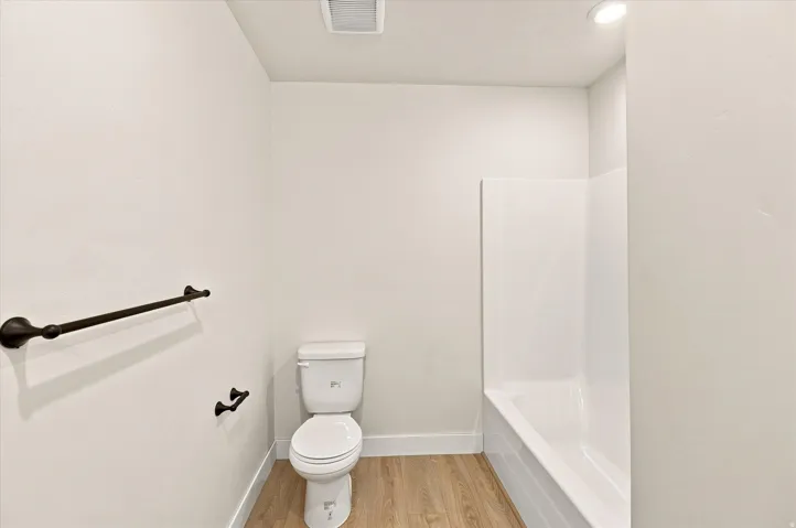 Bathroom featuring a white toilet, white bathtub with wall surround, wood-finish flooring, and dark-finish towel bars