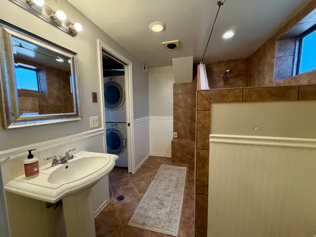 Pedestal sink with chrome faucet, framed mirror, built-in shower with tile surround, and stacked laundry appliances