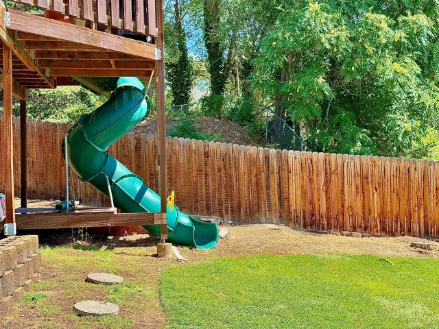Wooden deck structure with a spiral slide, complemented by a wood picket fence, a retaining wall, and a grassy lawn area