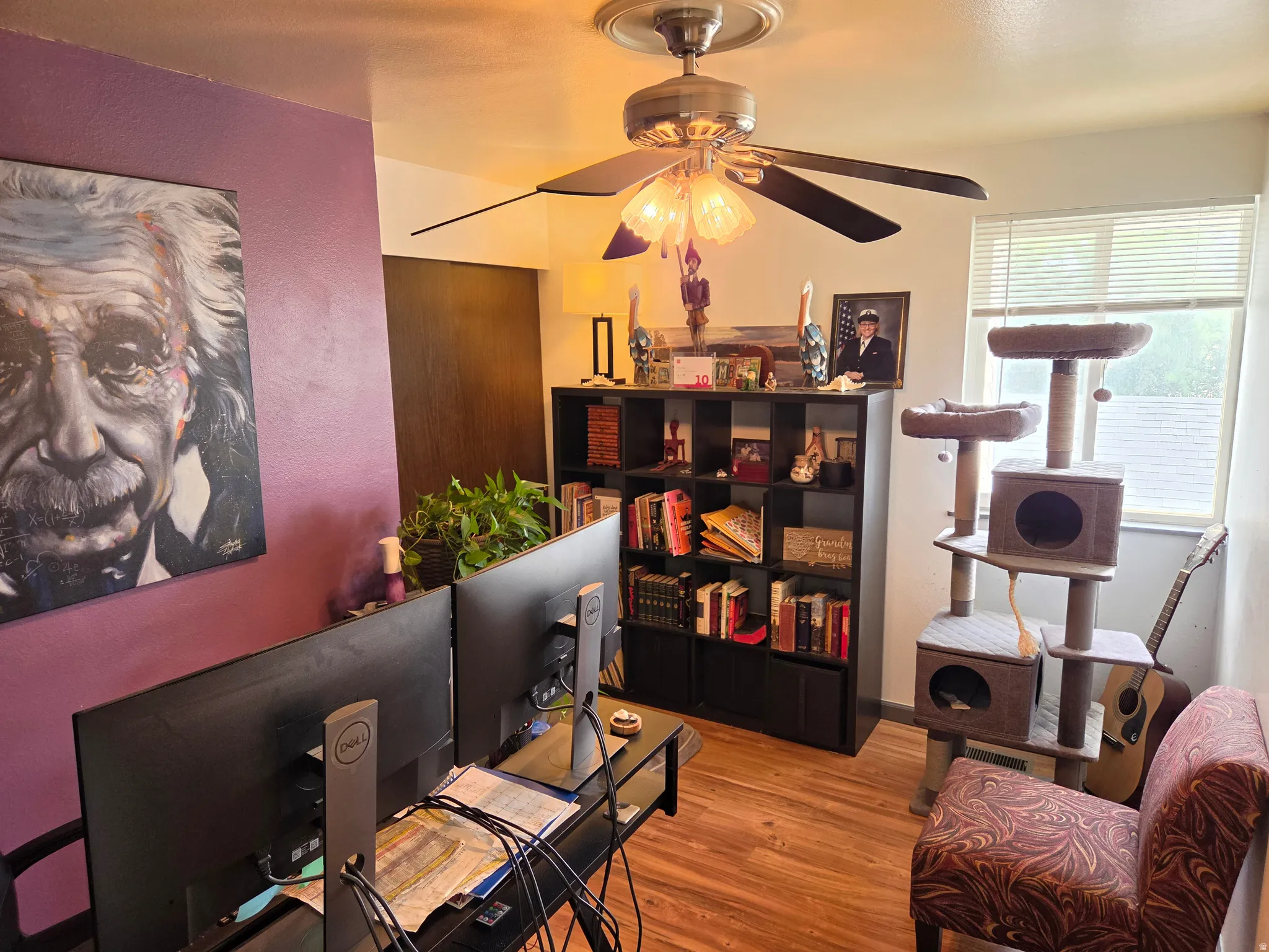 Wood-finish flooring, ceiling fan with light fixture, window with blinds, purple accent wall, and built-in shelving unit