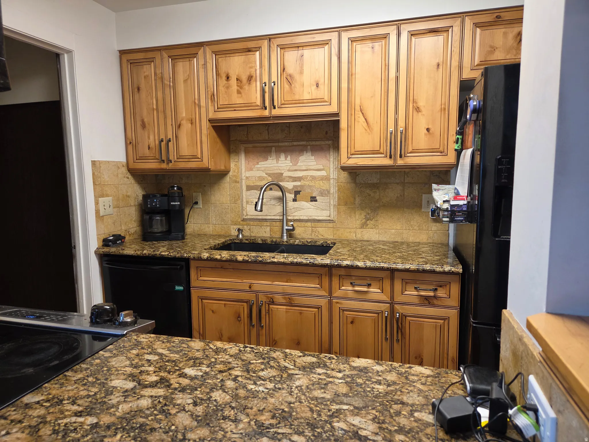 Wood-finish cabinetry with dark hardware, stone countertops, stone tile backsplash with a decorative insert, stainless steel faucet, and an undermount sink