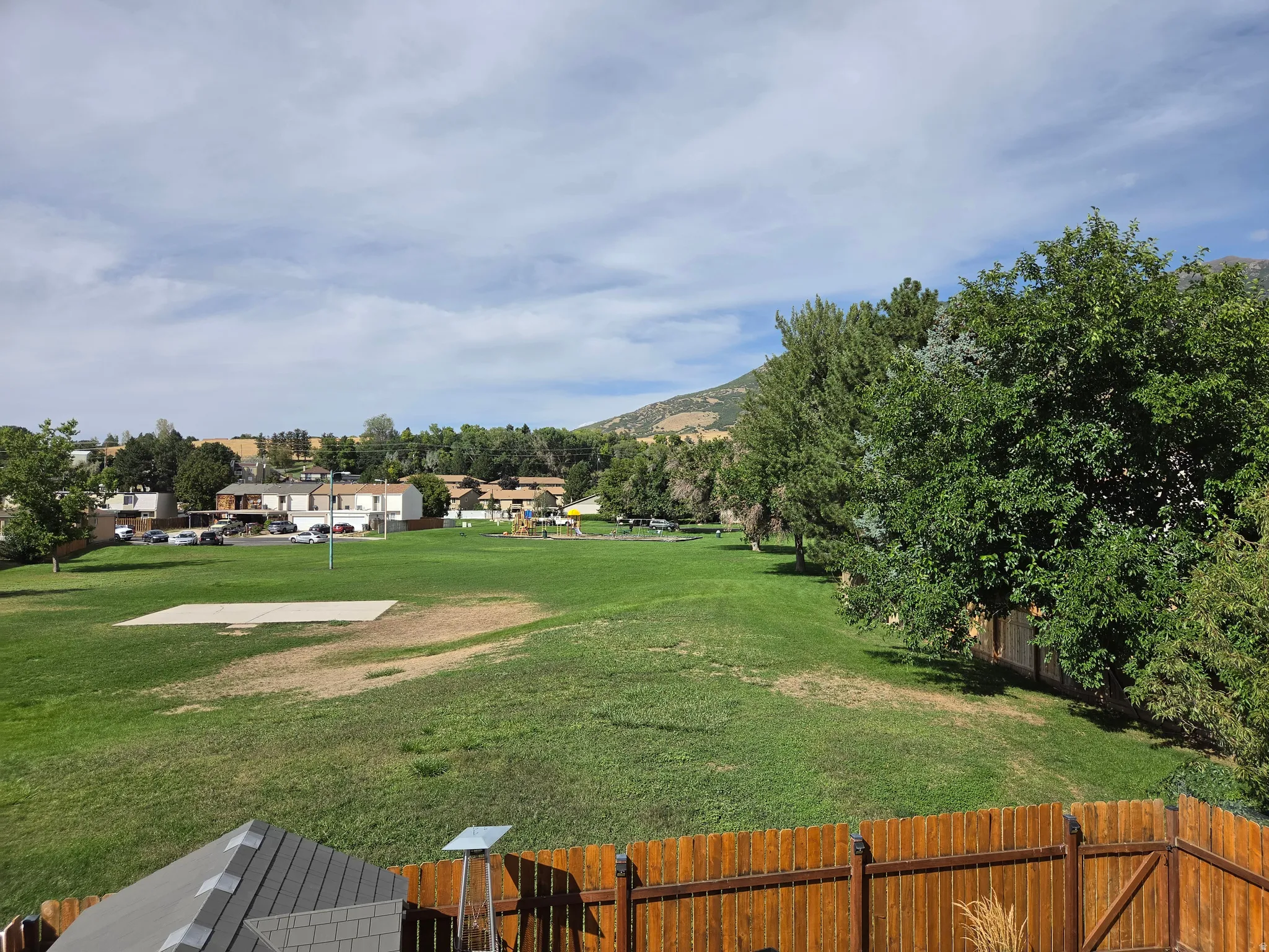 Expansive green space with distant residential structures, mature trees, and mountainous terrain under a partly cloudy sky