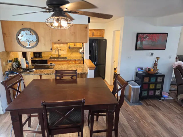 Open-concept dining area with wood-finish flooring, a ceiling fan with integrated lighting, and a pass-through kitchen featuring granite-style countertops and a tile backsplash