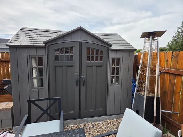 Gabled roof storage shed with gray shingle-style roofing, arched double doors with window panes, and two side windows
