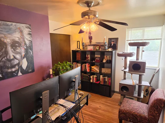Wood-finish flooring, ceiling fan with light fixture, window with blinds, purple accent wall, and built-in shelving unit