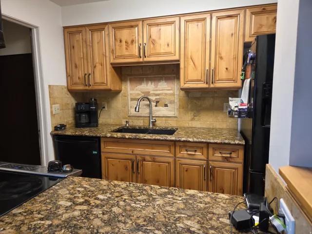 Wood-finish cabinetry with dark hardware, stone countertops, stone tile backsplash with a decorative insert, stainless steel faucet, and an undermount sink
