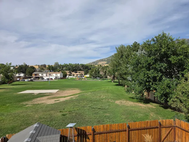 Expansive green space with distant residential structures, mature trees, and mountainous terrain under a partly cloudy sky